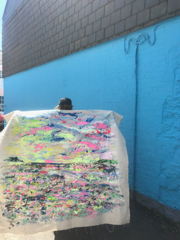 Contemporary Artist Chloe Tinsley holds out her canvas behind her like wings, a painting of Black Rock in the Falmouth Harbour, Cornwall in Greens and Pinks, Against a Bright Blue Wall in Flushing, Cornwall, green painting