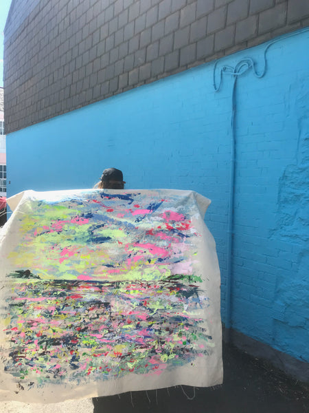 Contemporary Artist Chloe Tinsley holds out her canvas behind her like wings, a painting of Black Rock in the Falmouth Harbour, Cornwall in Greens and Pinks, Against a Bright Blue Wall in Flushing, Cornwall, green painting