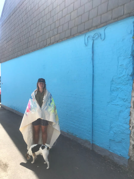 Contemporary Artist Chloe Tinsley is wrapped up in her canvas, a painting of Black Rock in the Falmouth Harbour, Cornwall in Greens and Pinks, Against a Bright Blue Wall in Flushing, Cornwall, a dog licks sniffs the ground in front of her