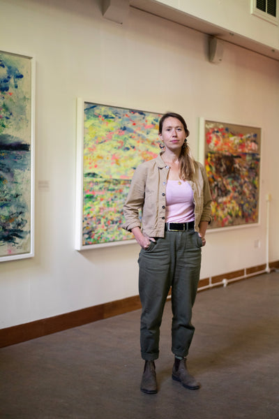 artist Chloe Tinsley stands in front of her Cornish Paintings at the exhibition at Trebah Gardens, the photo was taken by Kasia Murfet, Chloe wears Finisterre trousers and jacket, Blundstone tribe